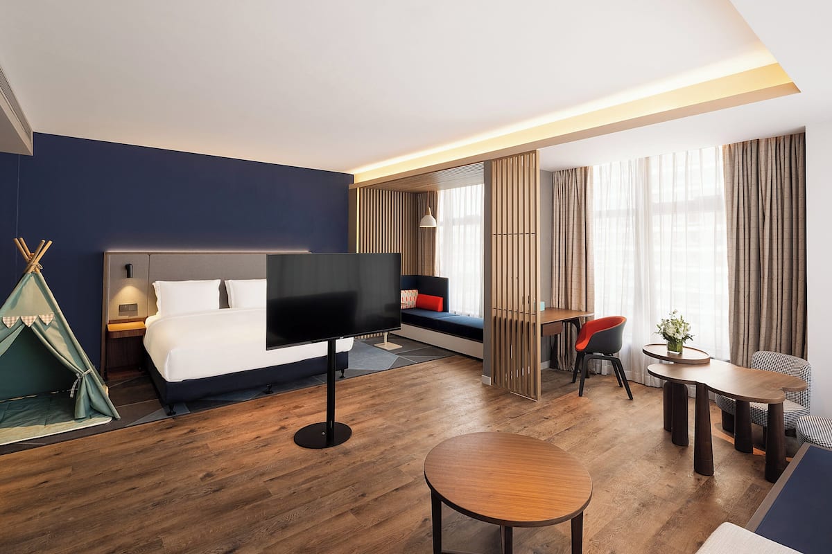 A hotel room with a bed, a TV, a dining table, a chair, a small table with a plant, and a tent-like structure.