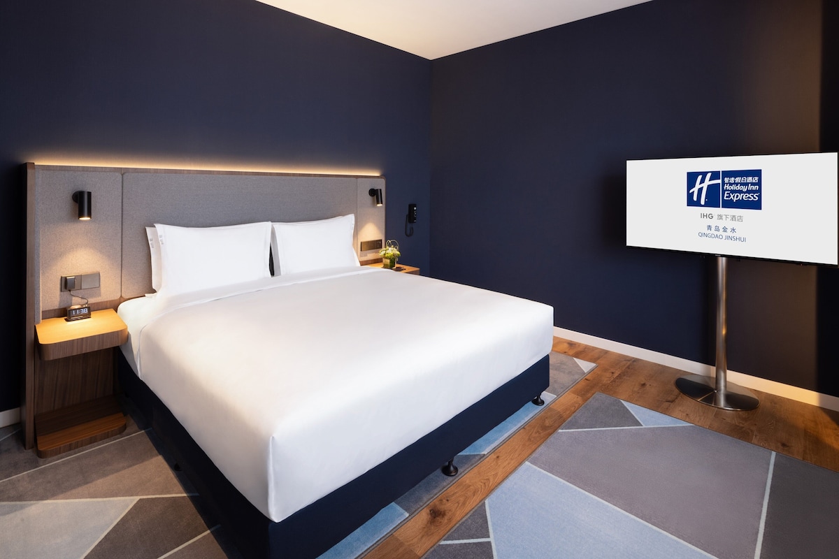 Une chambre d’hôtel moderne avec un grand lit, des tables de chevet avec des lampes, une petite table et un panneau indiquant qu’il s’agit d’un Holiday Inn Express.
