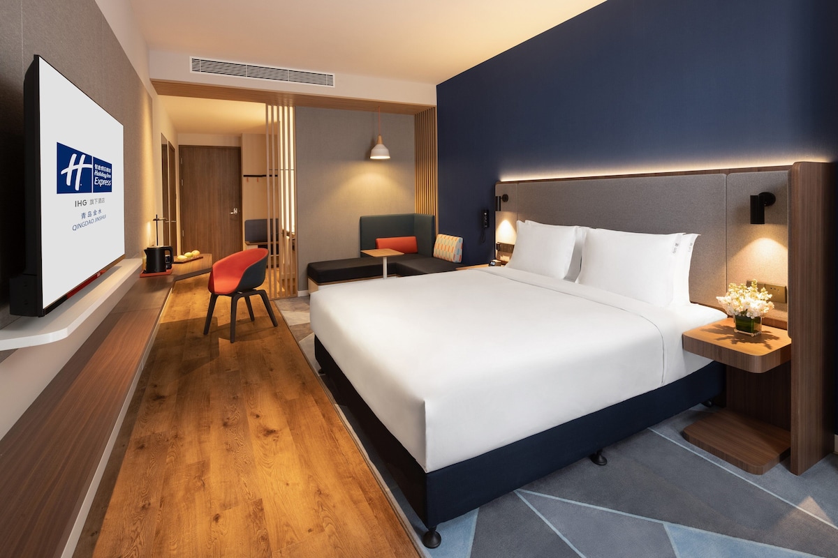 Une chambre d’hôtel moderne dotée d’un grand lit, de tables de chevet, d’une télévision et d’un coin salon avec un fauteuil et une table.
