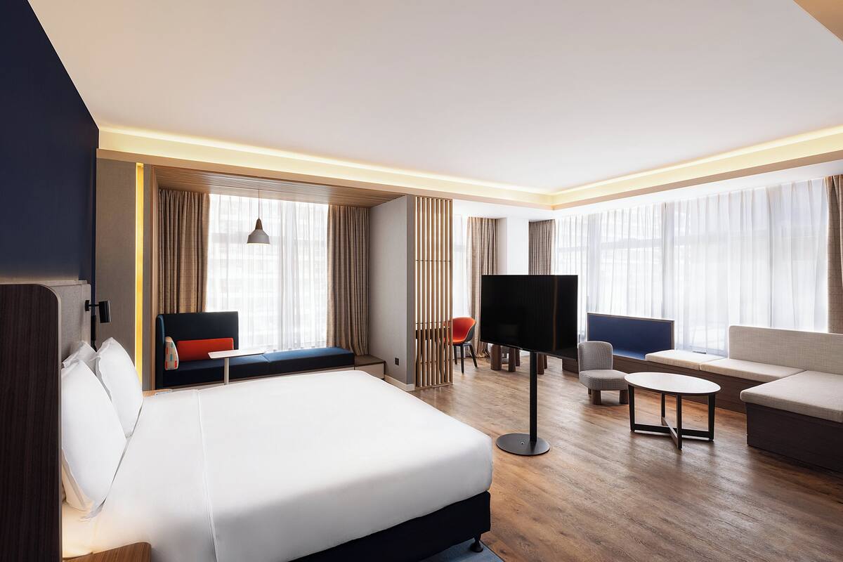 A hotel room with a bed, seating area, TV, and large windows.