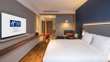 Una habitación de hotel moderna con una cama grande, un televisor en la pared, un escritorio con una silla y una zona de estar con sofá y mesa.