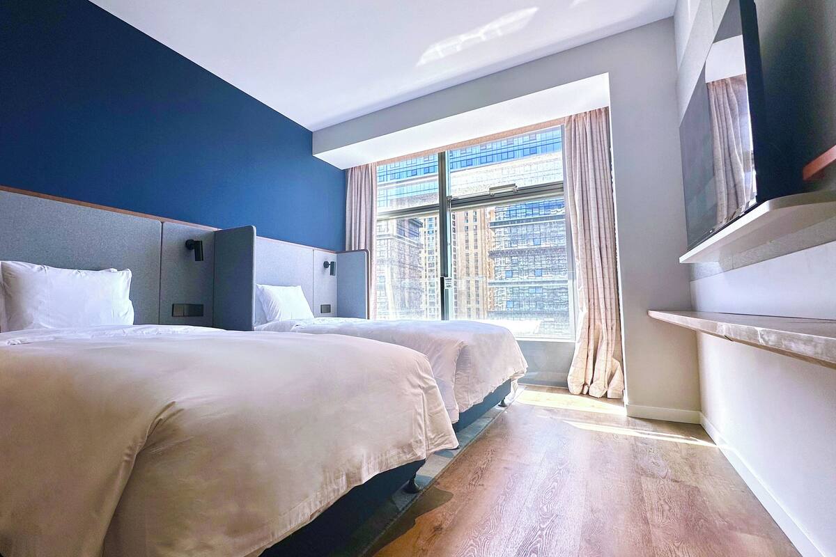 A hotel room with two beds, a large window with a city view, a flat-screen TV, and a wooden floor.