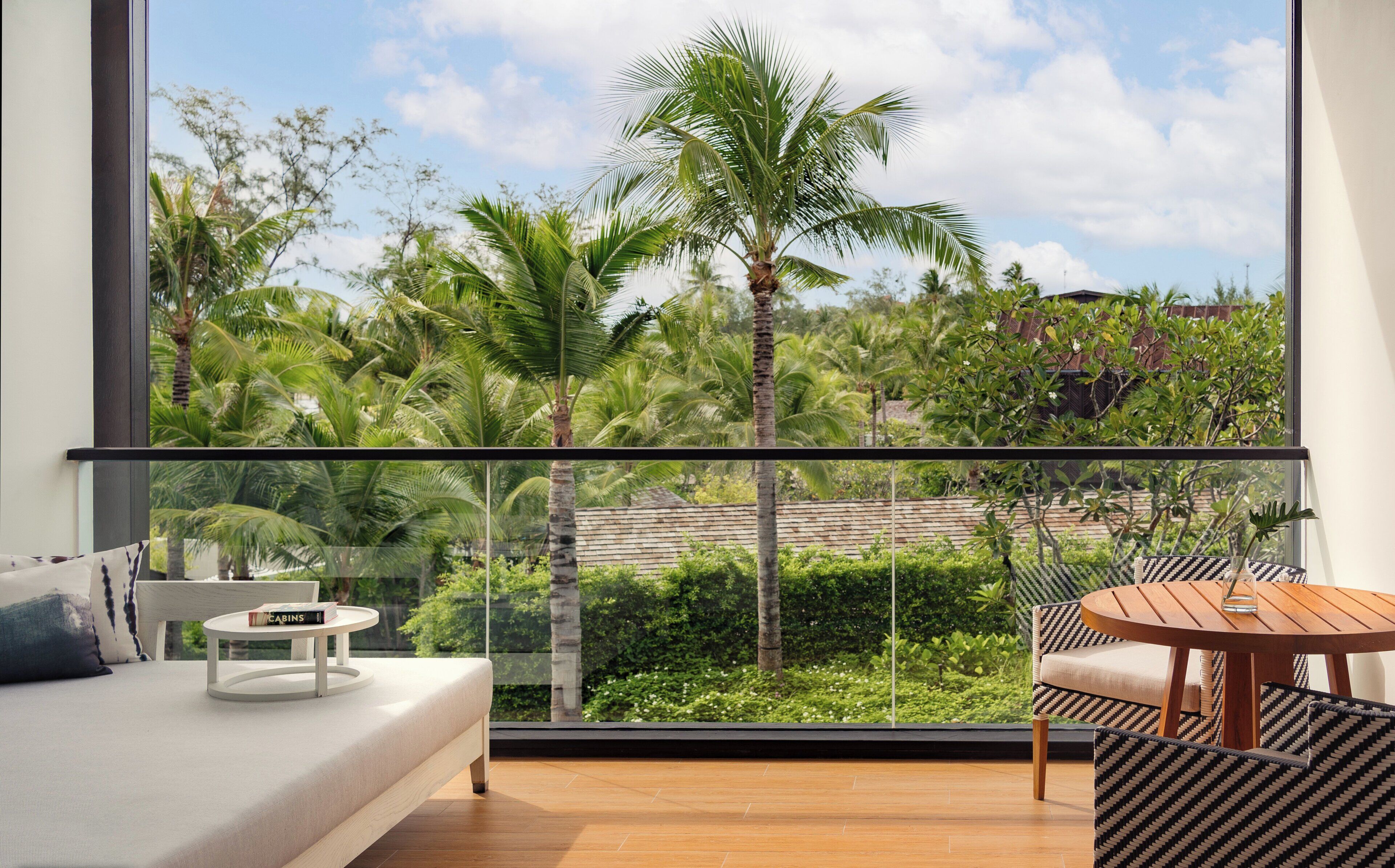 A balcony with a sofa, a small table, and a chair, overlooking a lush garden with palm trees and a clear sky.