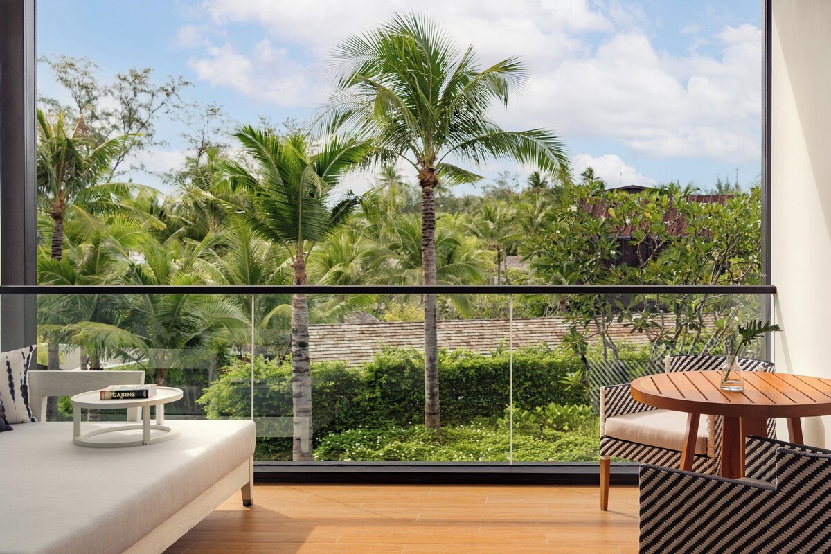 A balcony with a sofa and a small table, overlooking a lush garden with palm trees and a clear sky.