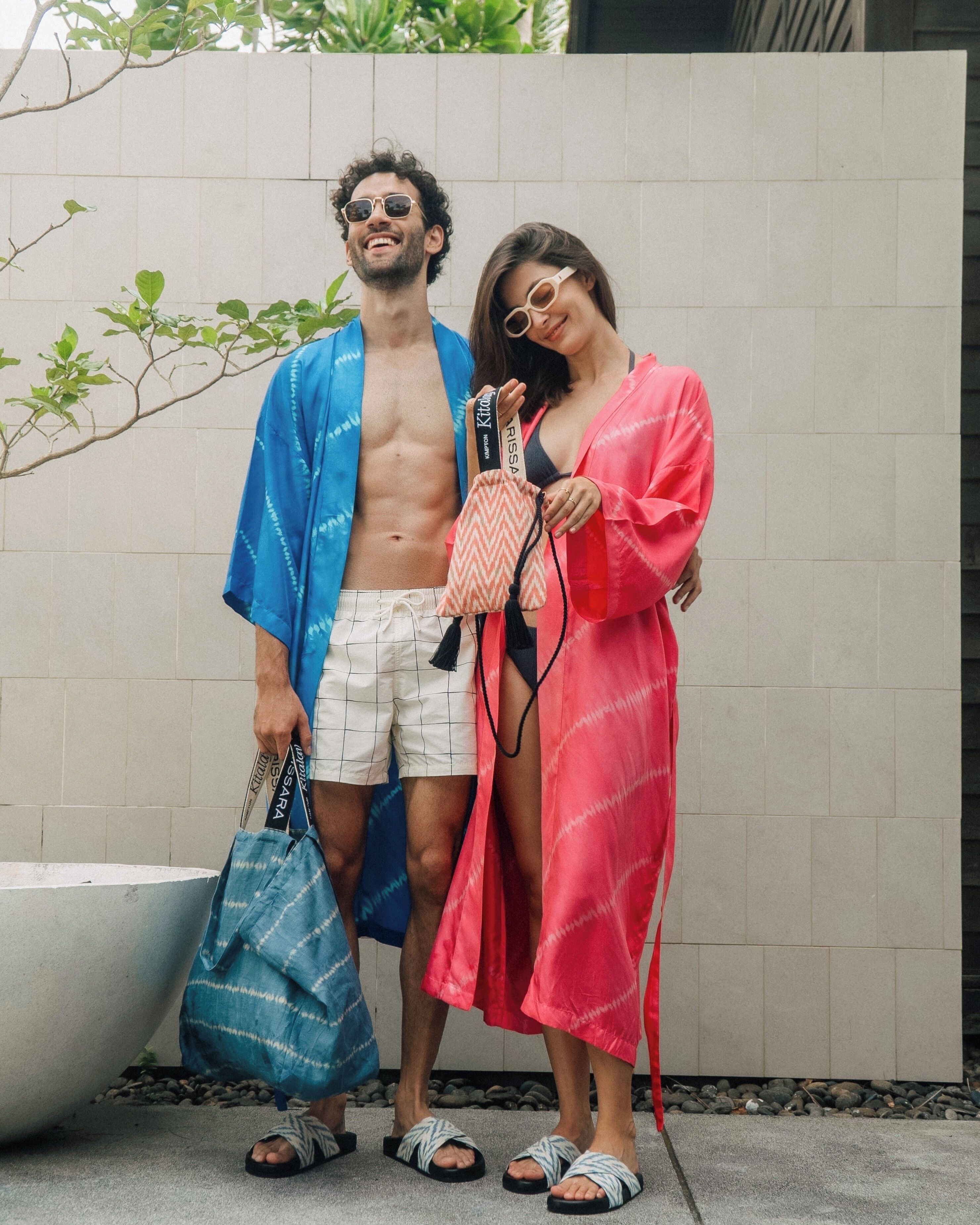 A man and a woman standing in front of a tiled wall, holding a striped bag and a small handbag.