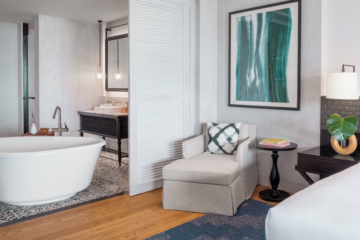 A modern hotel room with a large bathtub, a small armchair, a bedside table with a lamp, and a large window.