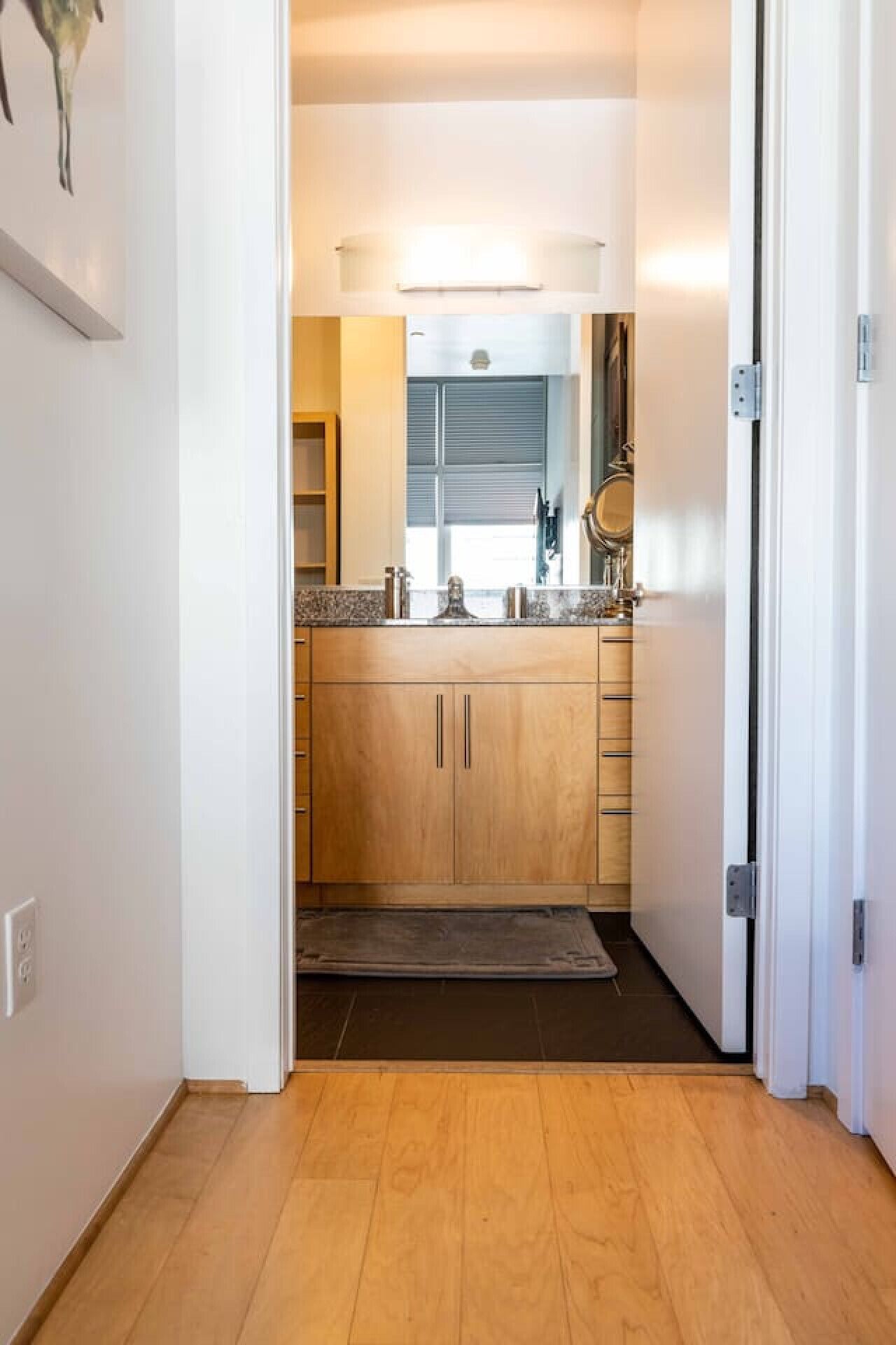Un couloir étroit mène à une cuisine équipée de meubles en bois, d’un plan de travail et d’une fenêtre avec des stores.
