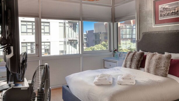 Une chambre d’hôtel moderne avec un grand lit, deux ventilateurs sur pied, un lustre et une vue sur la ville grâce à de grandes fenêtres.