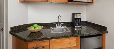 A compact kitchen with a granite countertop, a stainless steel sink, a coffee maker, and a microwave above the sink.