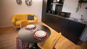 A compact living space with a dining table set for two, a yellow sofa, a kitchen area with dark cabinets, and a hanging plant. - Lemago Porto Apartments - POVEIROS 2B (Porto)
