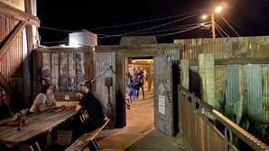A rustic outdoor dining area with wooden tables and benches, a metal water tank, and a wooden fence.