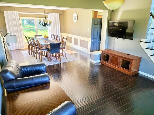 A spacious living area with a dark wood floor, a leather sofa, a dining table with chairs, a TV mounted on the wall, and a staircase leading to another level.