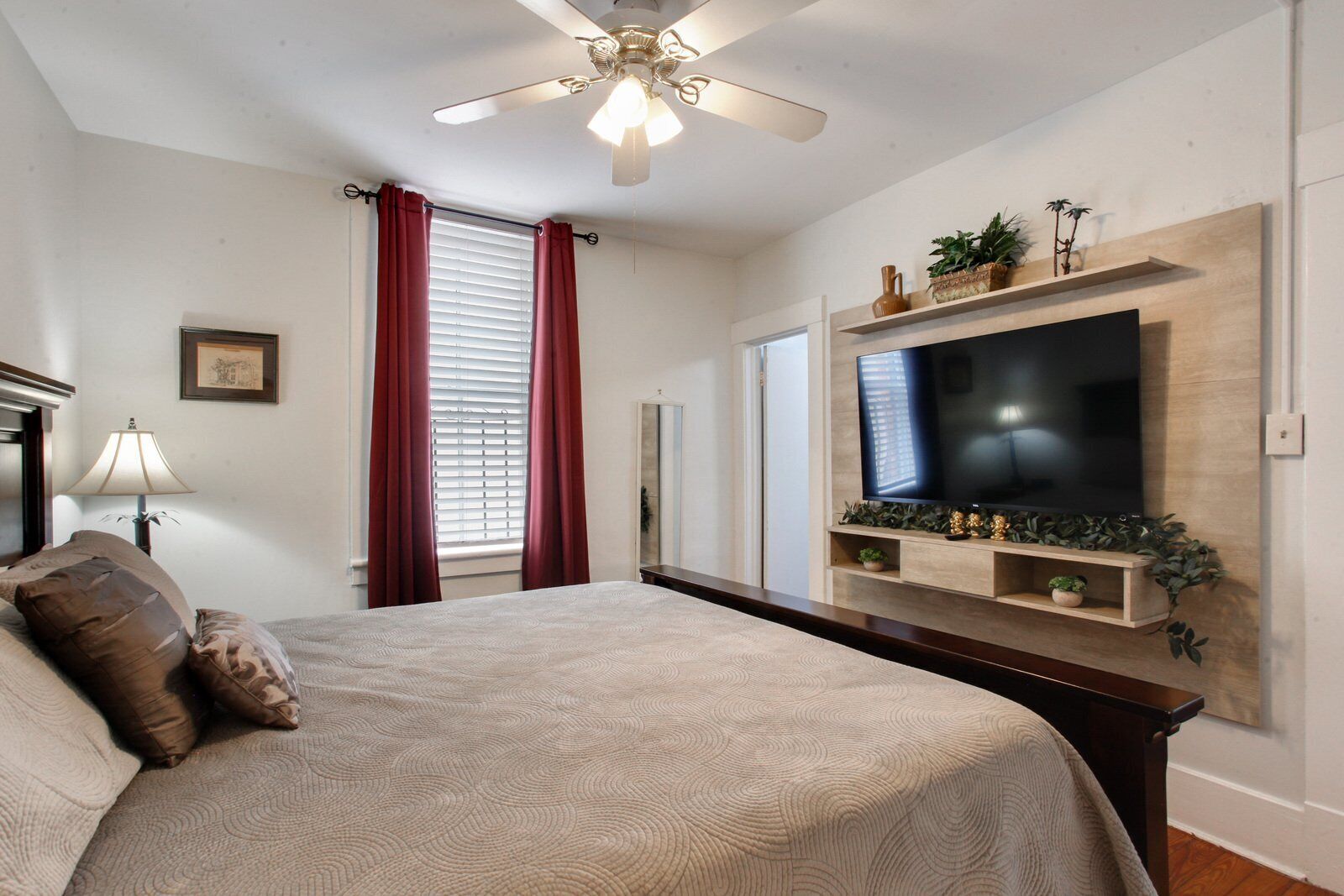 A bedroom with a bed, a ceiling fan, a TV mounted on the wall, and a window with curtains.
