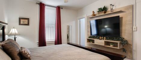 A bedroom with a bed, a ceiling fan, a TV mounted on the wall, and a window with curtains.