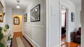 A hallway with white walls, wooden flooring, and a patterned rug. There are framed pictures on the walls and a mirror on the left. - Lincoln St Condo: Quiet Spot Near Night Life! (Savannah)