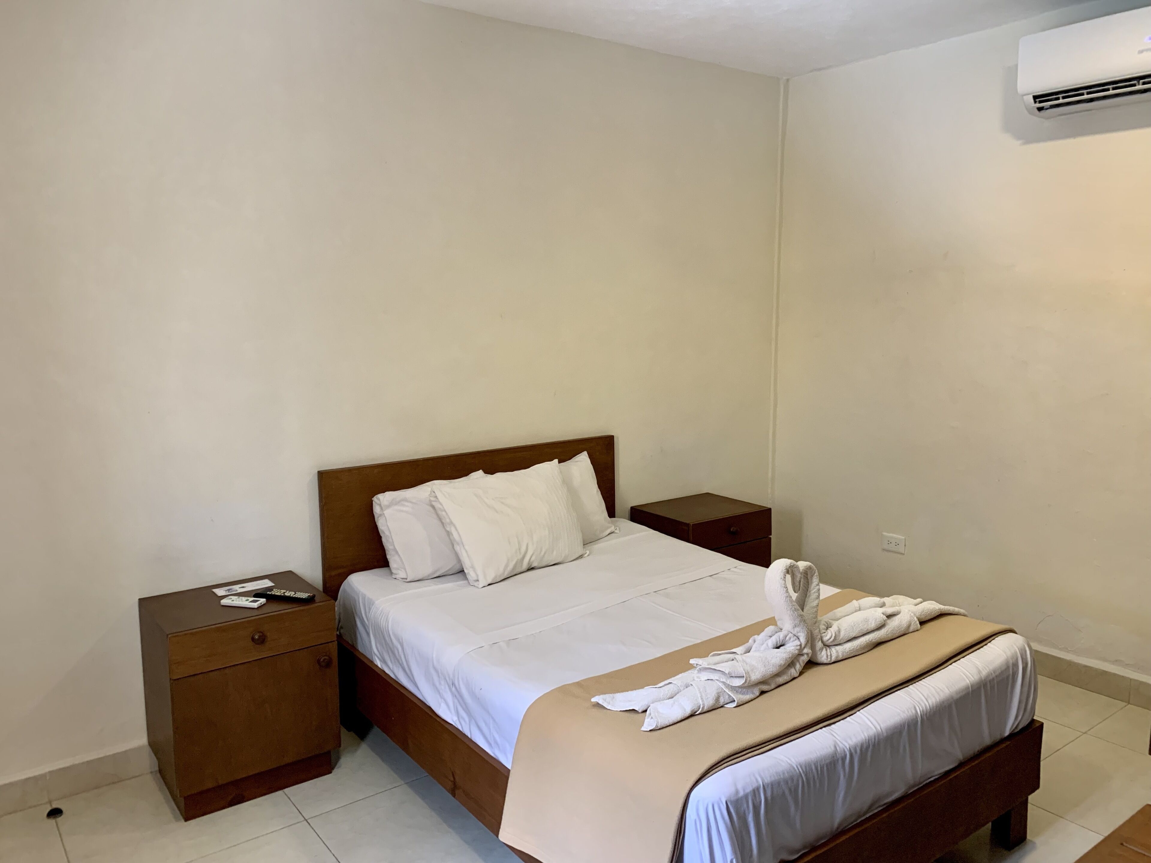 A neatly made bed with white linens and a towel arrangement, a wooden nightstand with a remote control, and a wall-mounted air conditioner.