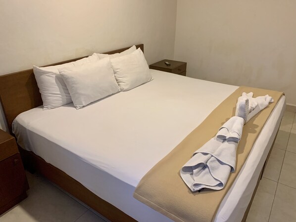 A neatly made bed with white linens and a beige blanket, a wooden headboard, and a small bedside table with a remote control.