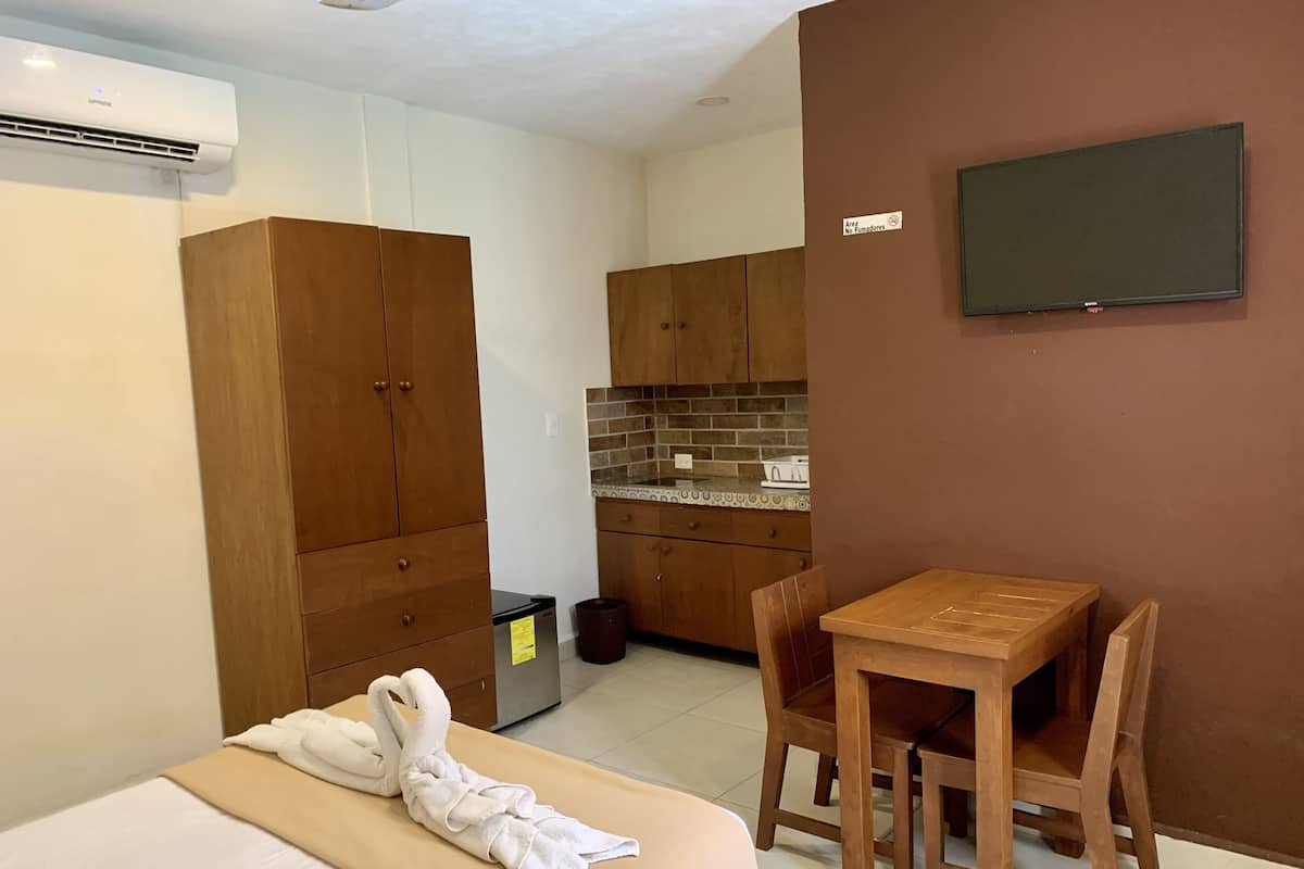 A hotel room with a bed, a wooden wardrobe, a small kitchen area, and a TV mounted on the wall.
