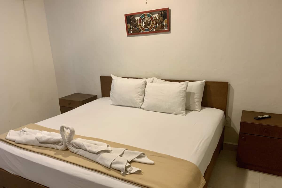 A neatly made bed with white linens and a beige throw blanket, a heart-shaped towel on the bed, and a small bedside table with a remote control.