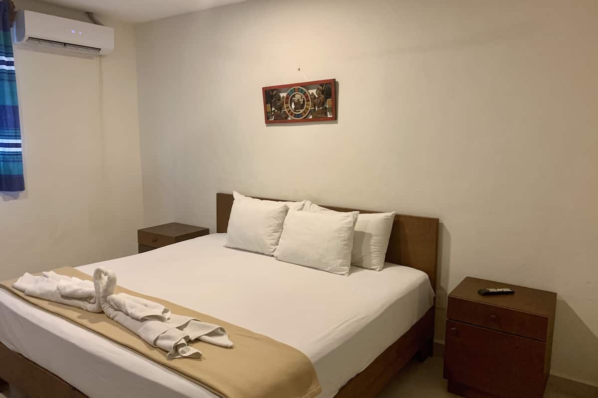 A neatly made bed with white linens and a beige blanket, a wooden headboard, a small bedside table with a remote control, and a wall-mounted artwork.