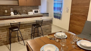 Een eetruimte met een houten tafel gedekt voor twee personen, een keuken met een magnetron en koffiezetapparaat, en een achterwand van baksteen.