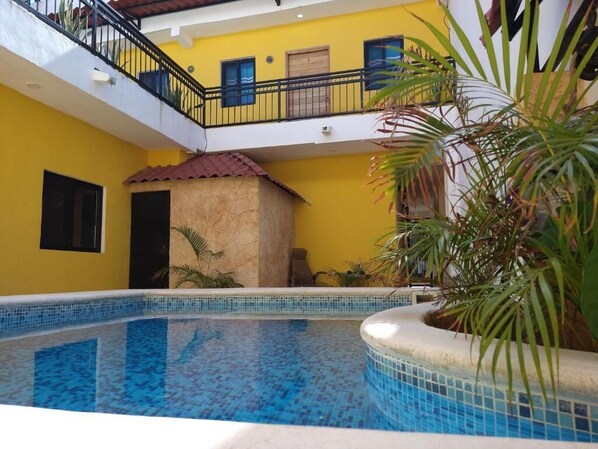 Una piscina con bordes de azulejos, un edificio amarillo con balcón y una planta de palmera.