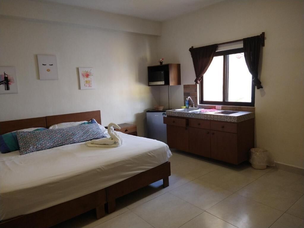 A bedroom with a bed, a bedside table, a small desk, a window with curtains, and a kitchenette with a sink and cabinets.