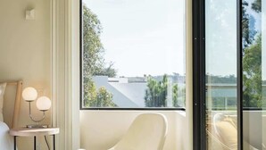 A modern hotel room with a large window, a white chair, a bedside table with lamps, and a bed.