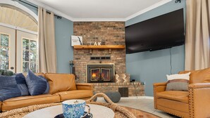 Ein Wohnzimmer mit Kamin aus Ziegelsteinen, Ledersofas, einem Flachbildfernseher und einem großen Fenster mit Vorhängen.