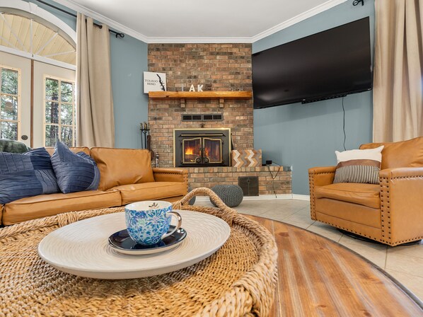 A living room with a brick fireplace, leather sofas, a flat-screen TV, and a large window with curtains.