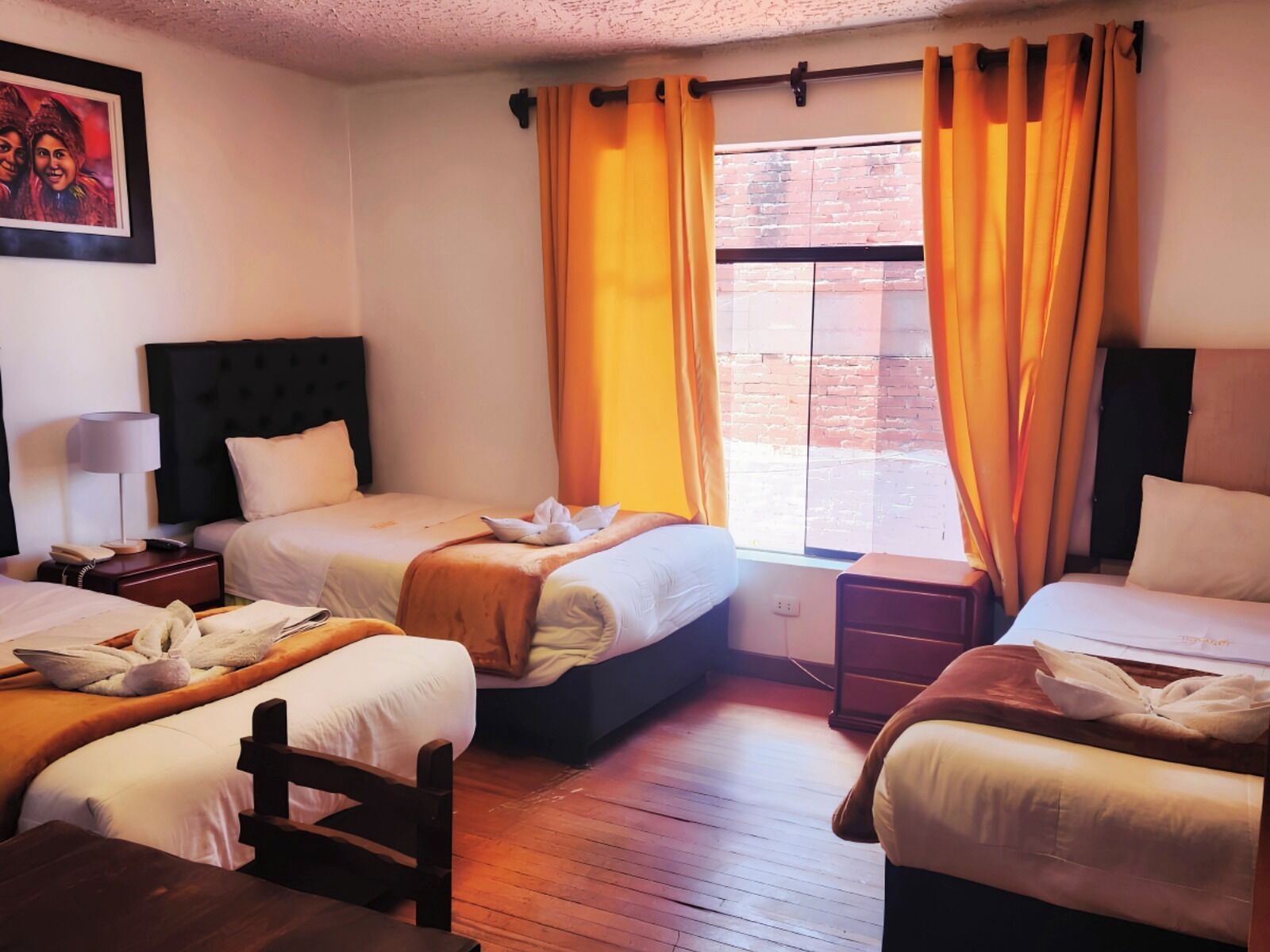 A hotel room with two beds, a wooden floor, a window with curtains, a bedside table with a lamp, and a framed picture on the wall.