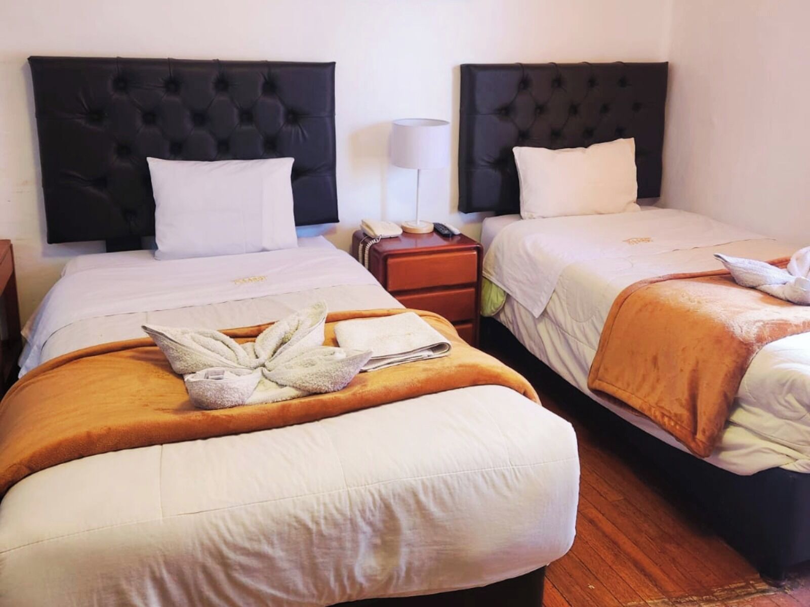A hotel room with two beds, each with a black headboard, white bedding, and a brown throw blanket. There is a bedside table with a lamp and a phone.