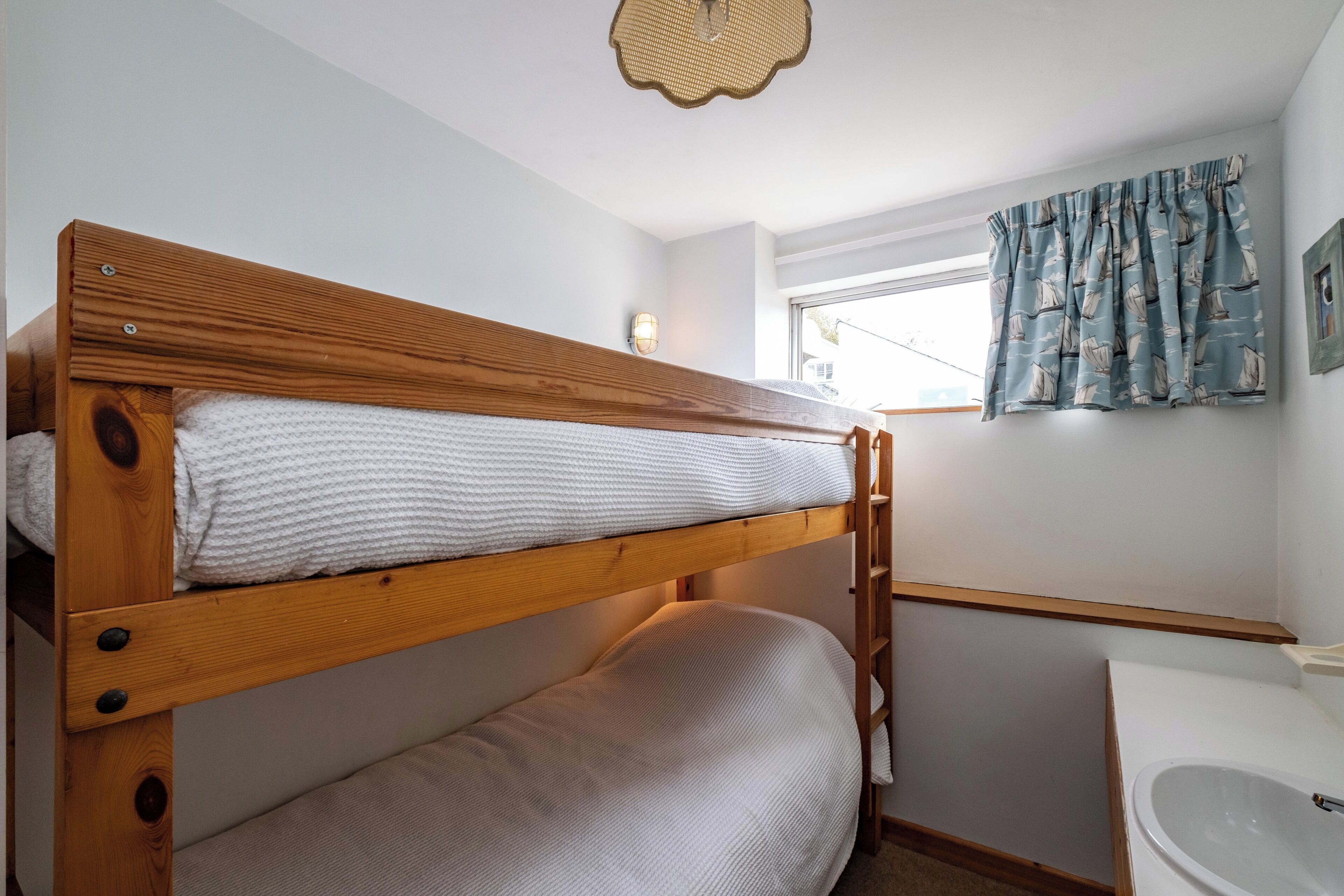 A bunk bed with a wooden frame and white bedding, a small window with curtains, and a white sink in a compact bathroom.