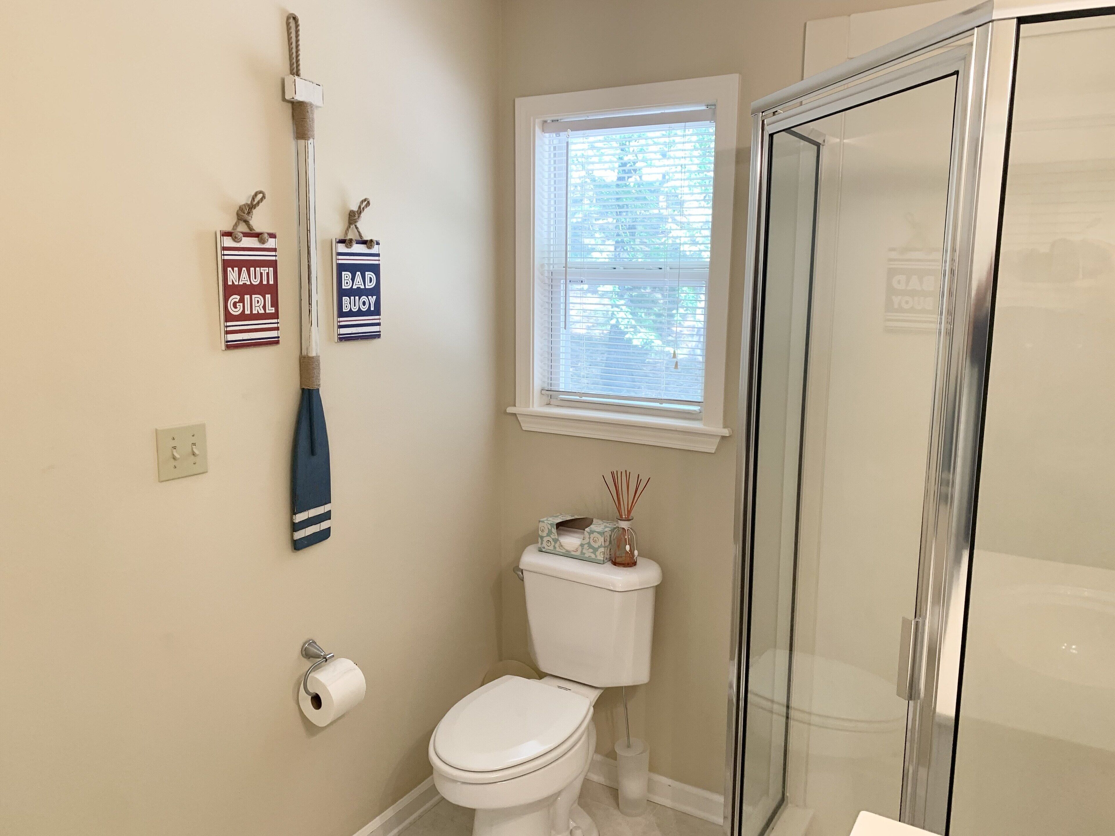 A bathroom with a toilet, a shower, a window with blinds, and two wall-mounted signs.