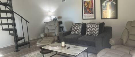 Una sala de estar de diseño moderno, con una escalera espiral negra, un sofá gris con cojines estampados, una mesa de centro de mármol y una obra de arte enmarcada en la pared.