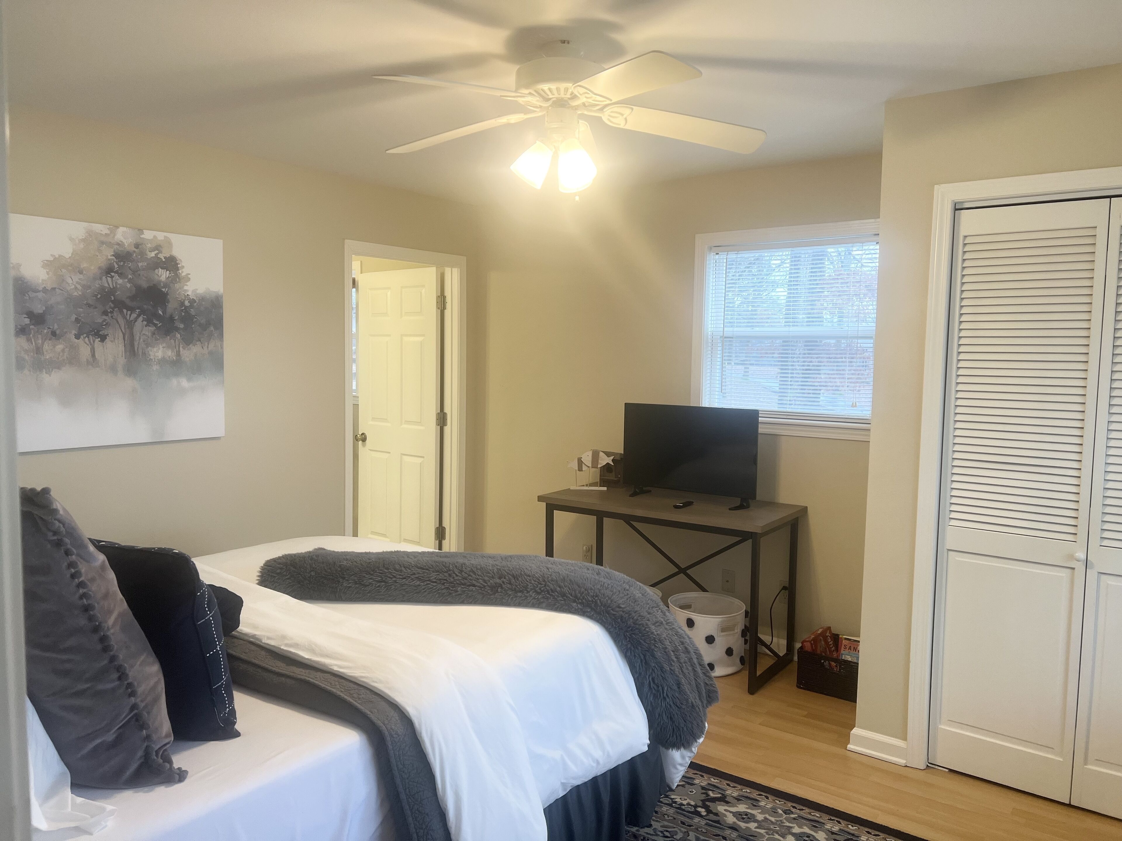 A bedroom with a bed, a ceiling fan, a TV on a small table, a window with blinds, and a door.