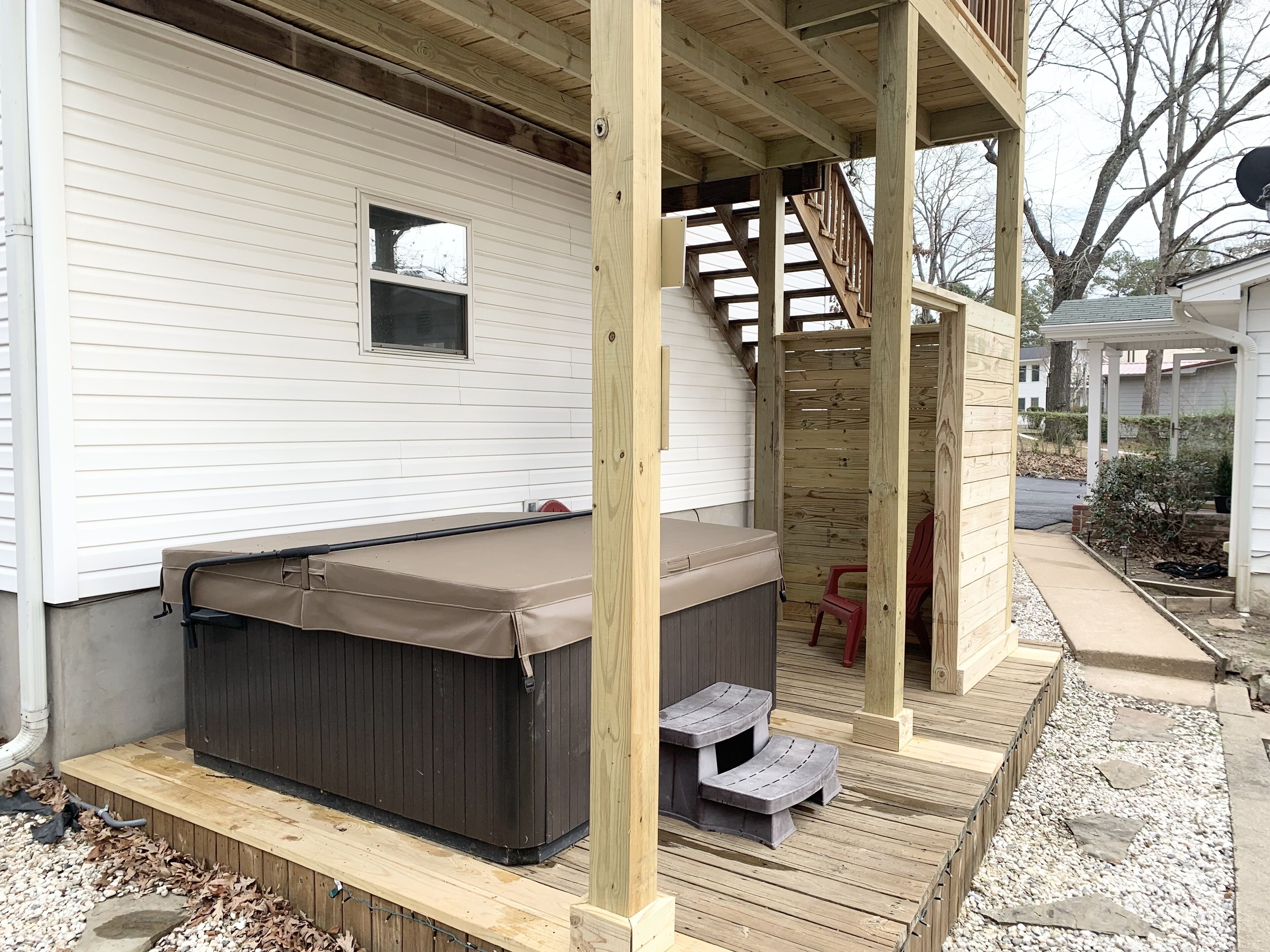 A hot tub on a wooden deck with steps leading up to it, a small window, and a covered area with a wooden railing.
