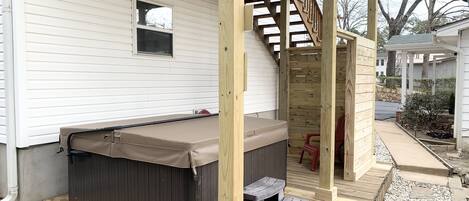 A hot tub on a wooden deck with steps leading up to it, a small window, and a covered area with a wooden railing.