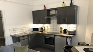 A compact kitchen with modern appliances, a dining area with a table and chairs, and a grey sofa with a green pillow.