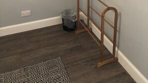 A corner of a room with a wooden hanger rack, a black metal rack, a small trash bin, and a patterned rug on the floor.