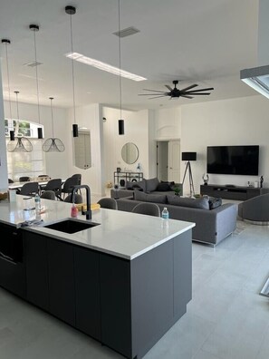 A modern kitchen with a central island, a dining area with chairs, a living room with a sofa and a TV, and a ceiling fan.