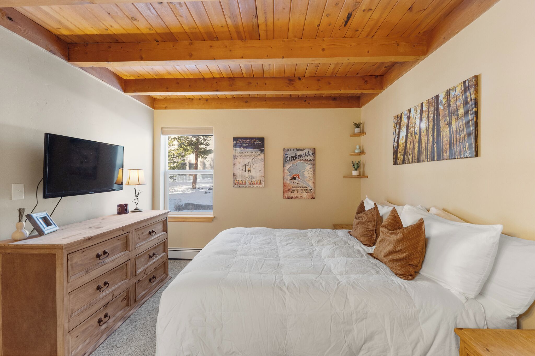 Un dormitorio con techo de madera, una cama con ropa de cama blanca, un televisor sobre un aparador de madera y dos carteles en la pared.