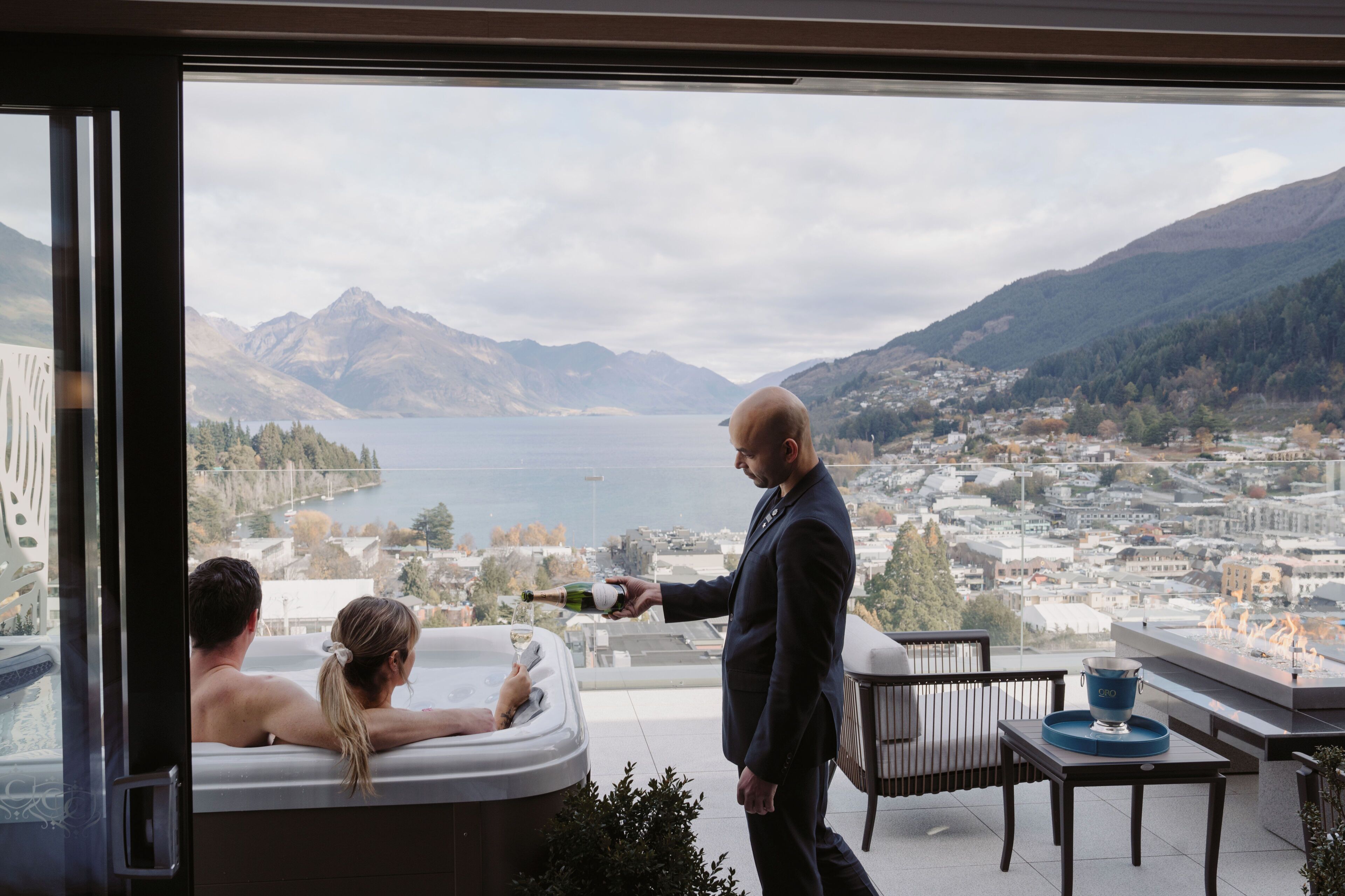 A man pouring champagne into a glass held by a woman sitting in a hot tub, with a scenic mountain and lake view in the background.