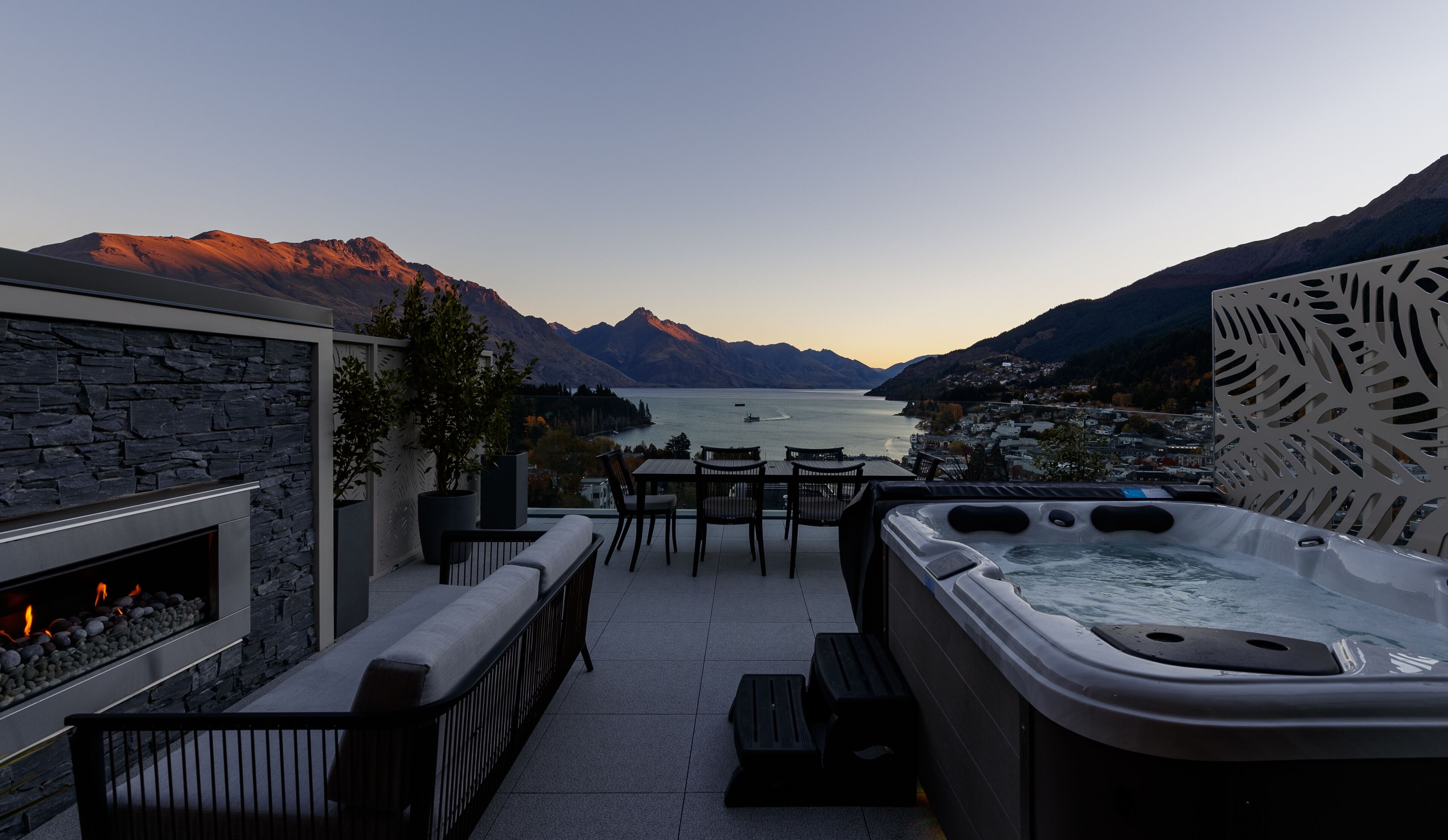 An outdoor terrace with a stone fireplace, seating area, and a hot tub overlooking a lake with mountains in the background.