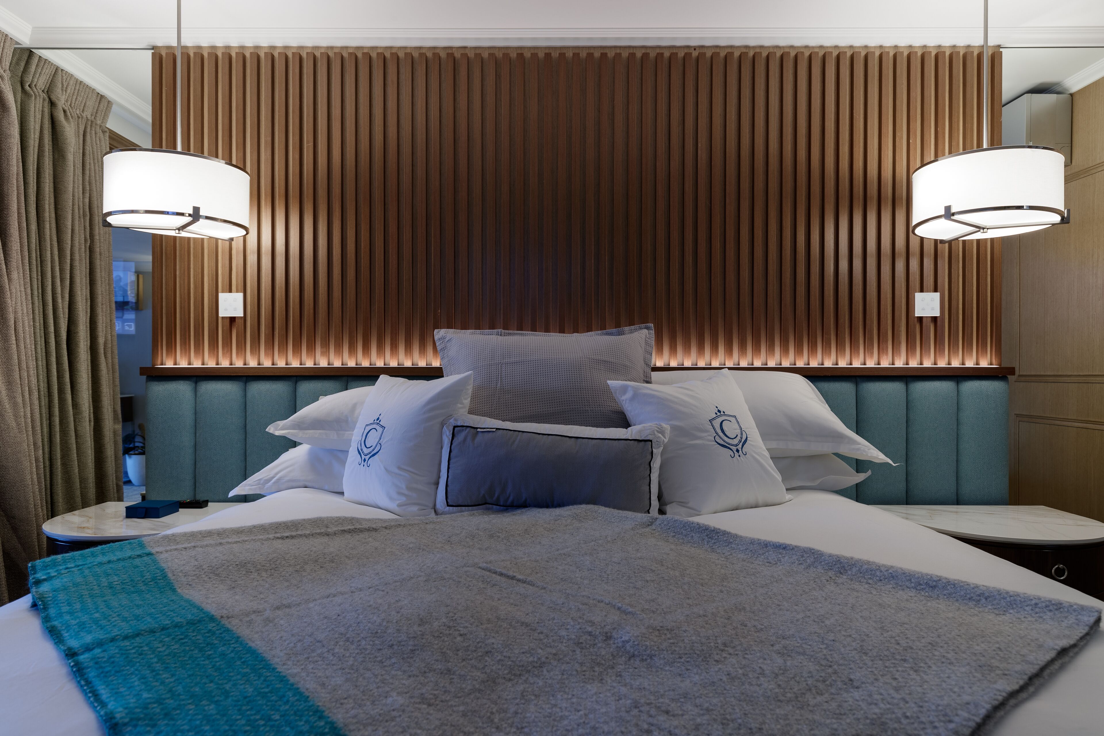 A modern hotel room with a large bed, two bedside tables, a headboard with vertical wooden slats, and two pendant lights.