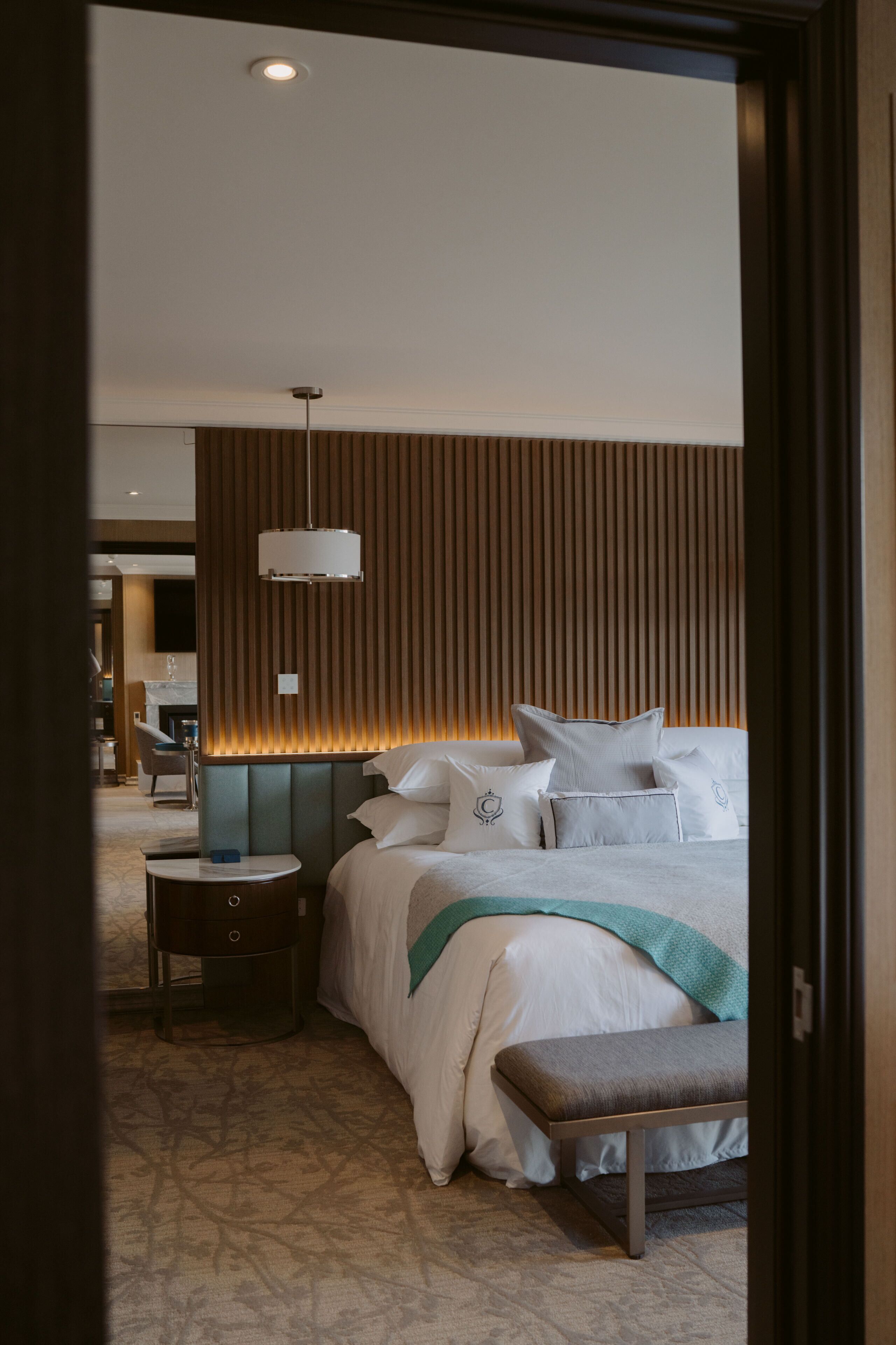 A modern hotel room with a large bed, a bedside table, a bench, and a wall-mounted light fixture.