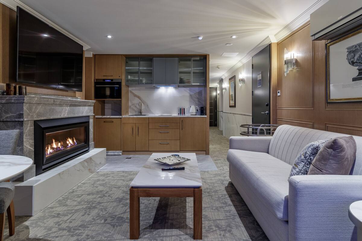 Een moderne woonkamer met een open haard, een bank, een salontafel en een keukenhoek met houten kastjes en een marmeren aanrecht.