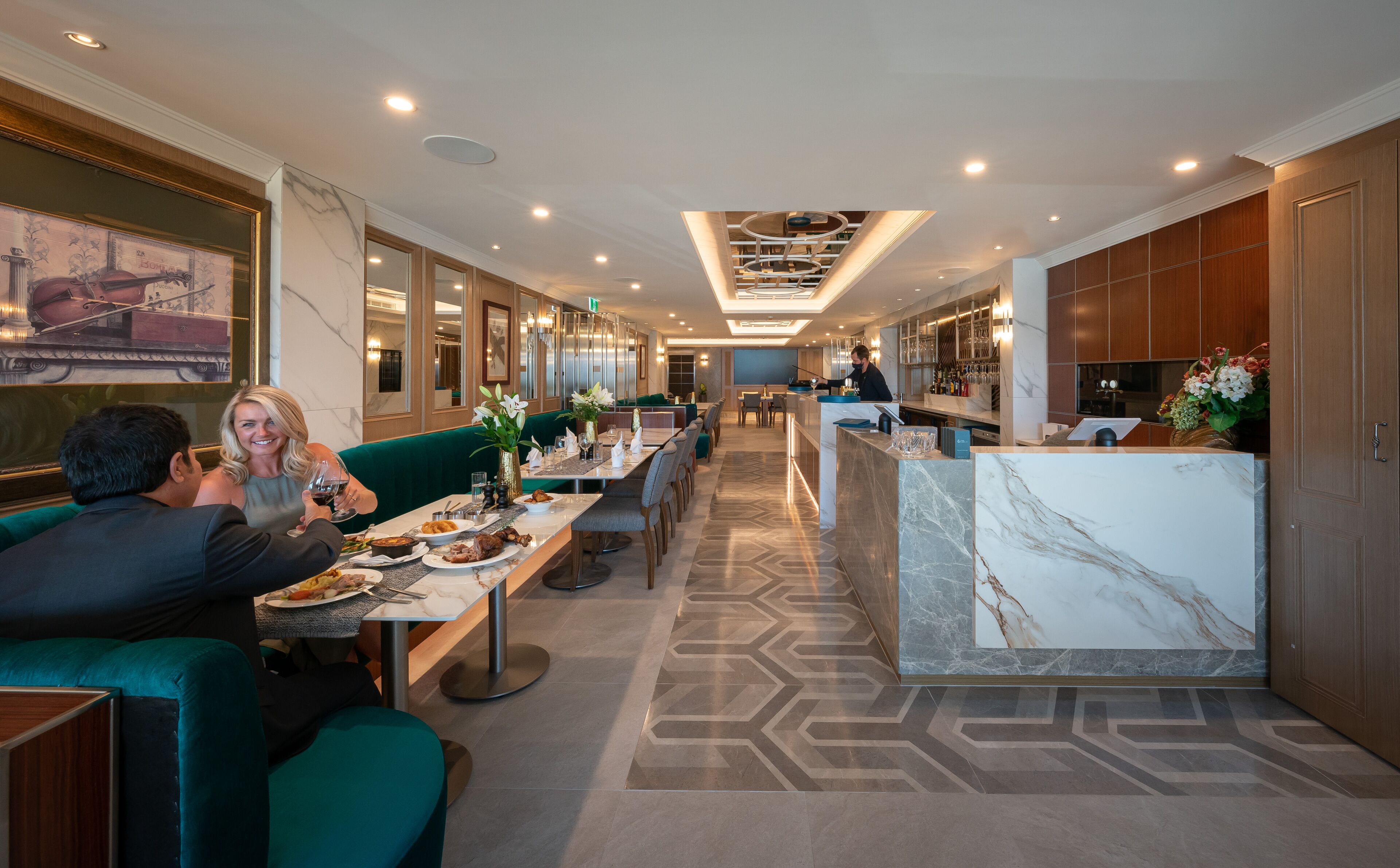 A modern hotel dining area with green seating, marble countertops, and a bar area with a bartender.