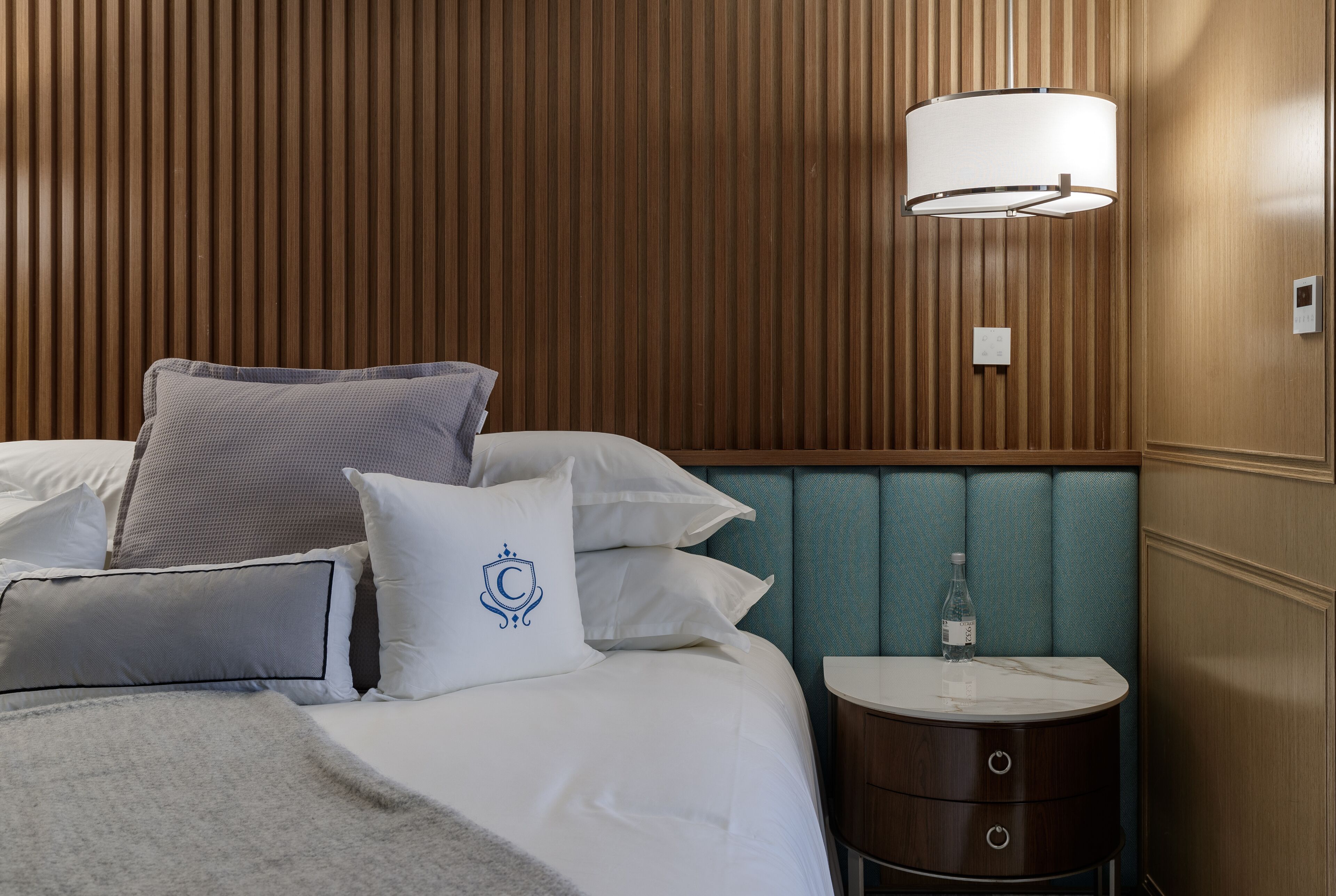 Una cama bien hecha con sábanas blancas y varios almohadones, una mesita de noche con una botella, y una lámpara fijada a la pared.