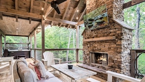 A covered outdoor area with a stone fireplace, seating, and a view of the forest. - Generational Cabin Near Gatlinburg & Dollywood (Sevierville)